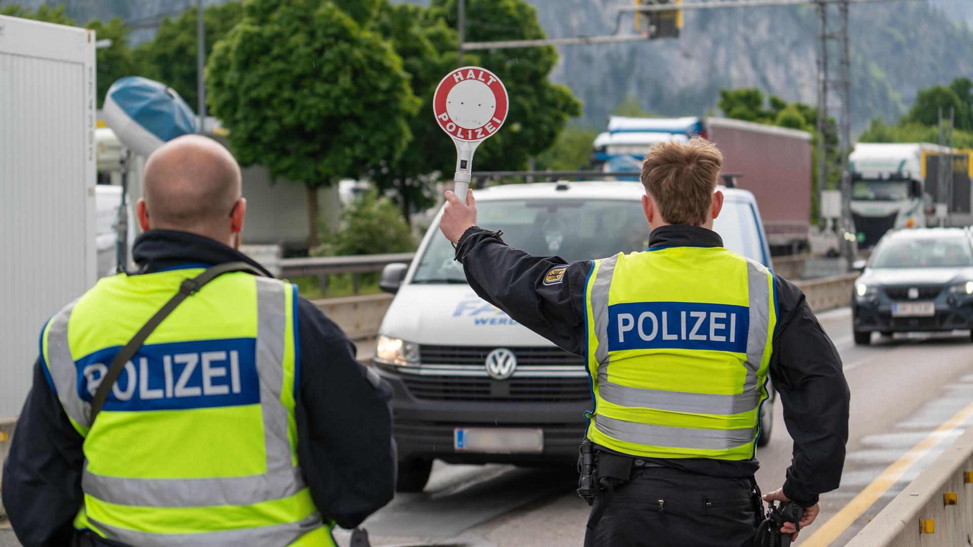 Ein Polizist stoppt ein Fahrzeug auf der Autobahn mit einer Polizeikelle mit der Aufschrift: "Halt Polizei". Neben ihm steht ein anderer Polizist.
