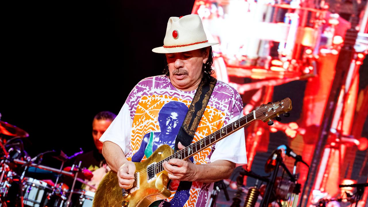 Carlos Santana steht auf der Bühne und spielt Gitarre. Er trägt einen weißen Hut und ein buntes Oberteil. 