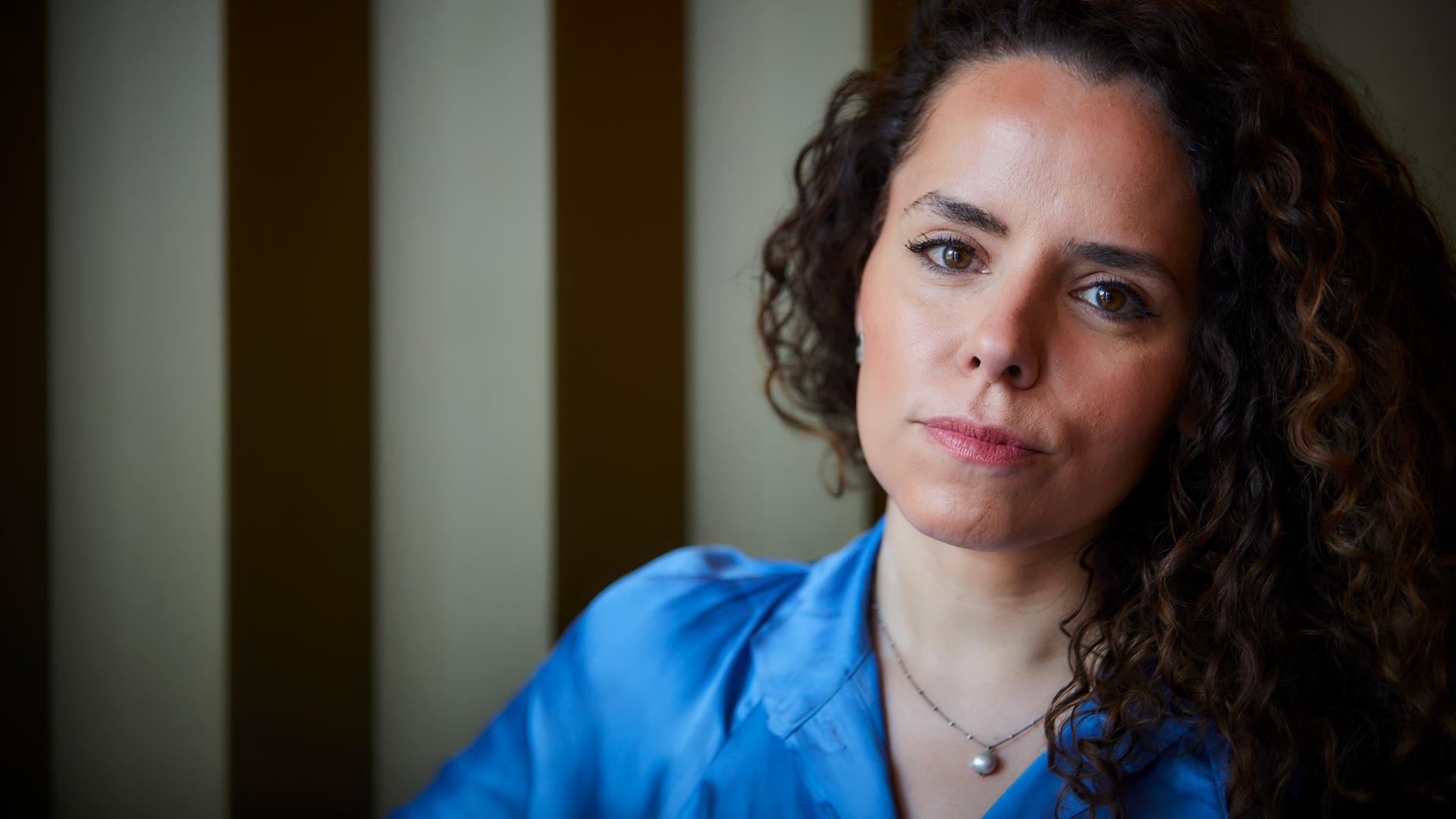 Portrait der Journalistin Alena Jabarine. Sie trägt eine helle blaue Bluse. Ihre langen, lockigen Haare liegen auf ihrer linken Schulter. Sie blickt mit entschlossenem Blick in die Kamera.